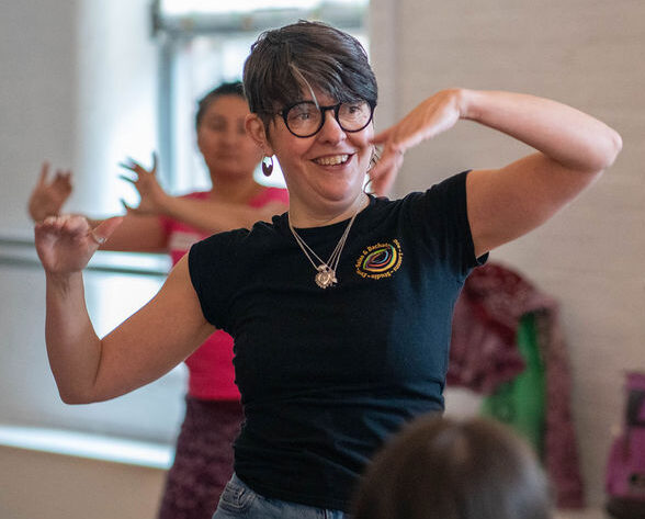 A middle-aged slender female session leader with short dark hair, wearing glasses with a black frame, silver jewellery, and a black T-shirt with short sleeves, a huge warm smile on her face, demonstrates a dance move with her arms in the IMPACD studio.