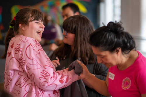 A young lady with brown hair in a patterned pink jumper and a huge beaming smile holds hands and dances with other young ladies in an inclusive movement session at the IMPACD studio.