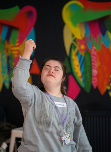 A young brown-haired lady with Down’s syndrome, wearing a grey jumper and with a serene expression on her face, lifts her right fist to the heavens, with colourful wings adorning the back wall of the IMPACD studio seemingly coming out of her back like an angel’s.