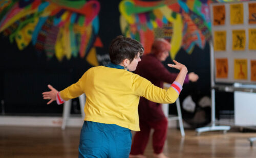 In the IMPACD studio, a middle-aged slender female session leader with short dark hair, sporting a yellow cardigan over a blue jumpsuit, faces a participant, wearing a red outfit and hair styled in a red bob cut, and moves her arms in the float motion from the IMP tool.