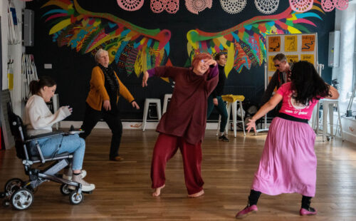 In the IMPACD studio, a tall bare-footed middle-aged female participant in a red outfit and her hair styled in a red bob cut, moves her arms in the float motion from the IMP tool surrounded by a diverse group of dancers of all ages, including a wheelchair user.