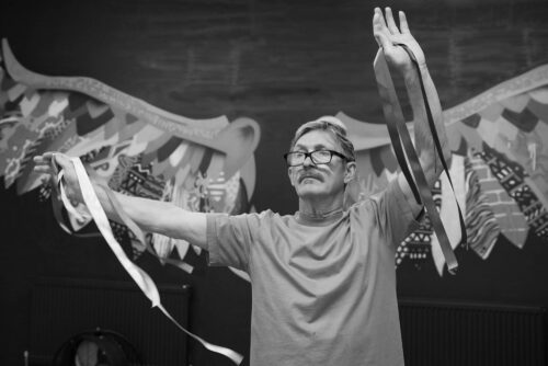 An older gentleman with a moustache and short salt-and-pepper hair, wearing a short-sleeved T-shirt and glasses with a black frame, stands tall and calmly holds ribbons at arms’ length in both hands in front of the beautiful patterned wings that adorn one of the walls in the IMPACD studio.