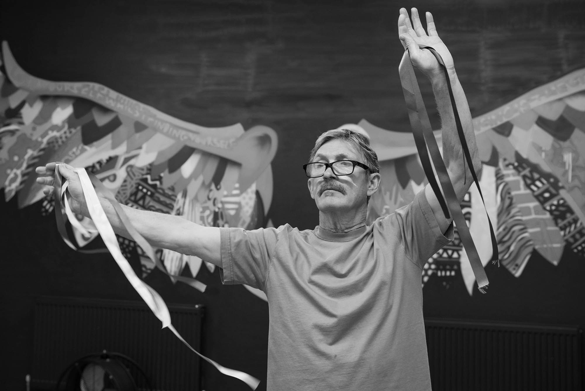 An older gentleman with a moustache and short salt-and-pepper hair, wearing a short-sleeved T-shirt and glasses with a black frame, stands tall and calmly holds ribbons at arms’ length in both hands in front of the beautiful patterned wings that adorn one of the walls in the IMPACD studio.