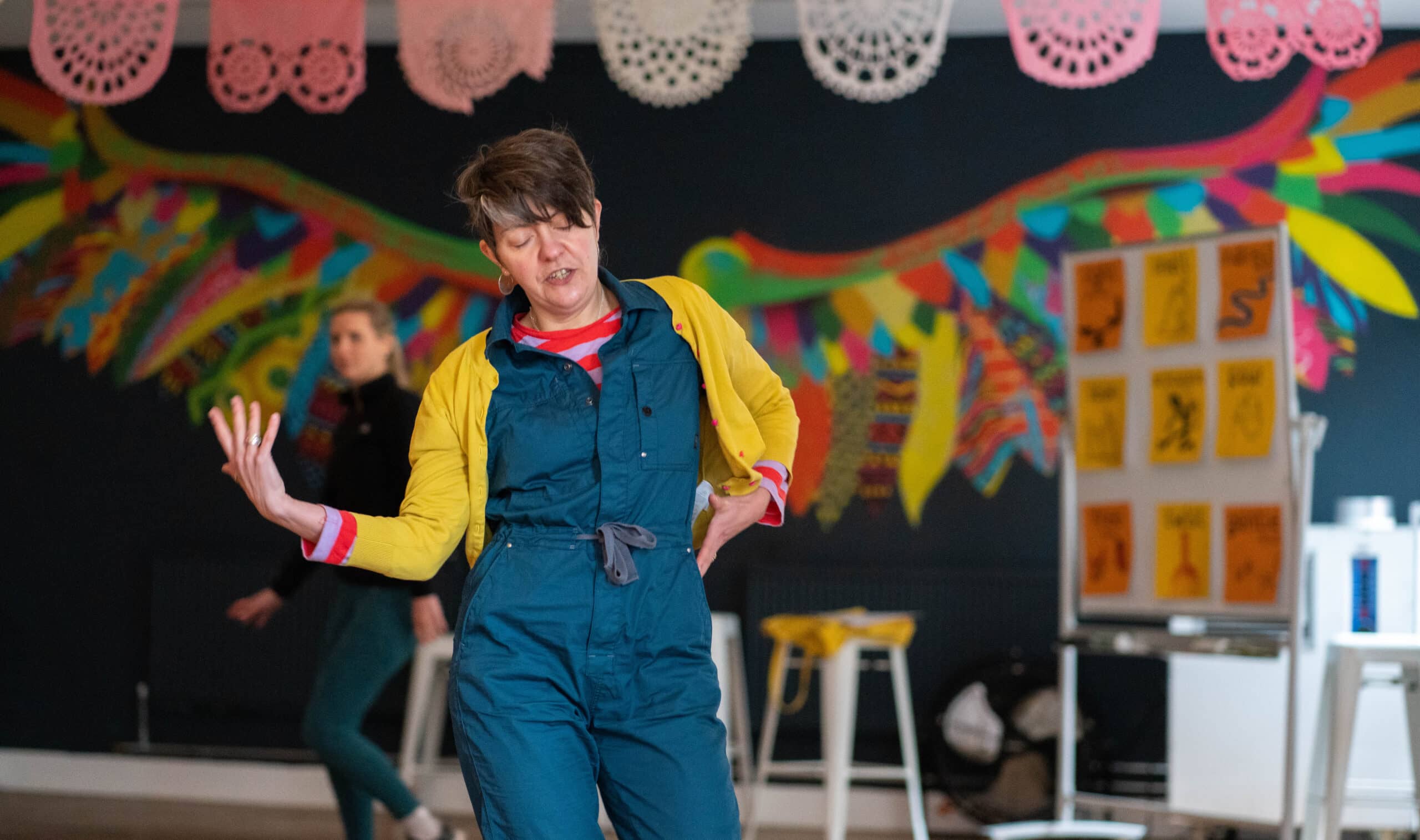 A middle-aged slender female session leader with short dark hair, wearing a blue jumpsuit and yellow cardigan, eyes closed and serene-looking, moves her arms pensively. Beautiful wings which adorn the back wall of the IMPACD studio seem to come out of her back like a bird in flight.