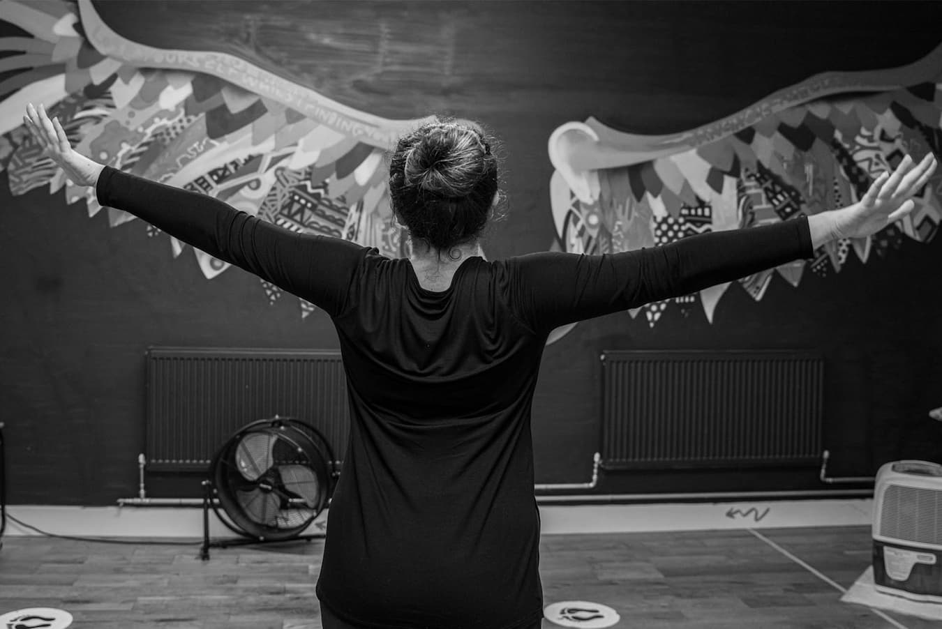 A slender lady with her hair in a bun, wearing a black long-sleeved top and her arms opened like a bird, faces away towards a wall in the IMPACD studio which is adorned with beautiful patterned wings that span as wide as her arms’ reach.